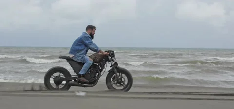 Bearded man in jeans rides old cafe racer motorcycle on sandy beach near sea Stock Footage 140624488