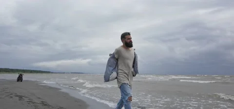 Bearded man in jeans on sandy beach near sea in slow motion, takes off jacket Stock Footage 140624772