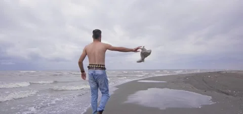 Bearded man in jeans on sandy beach near sea in slow motion, takes off shirt Stock Footage 140624779