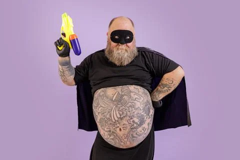 Bearded man with large tattooed abdomen in carnival costume holds toy blaster on Stock Photos