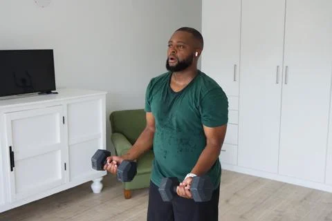 Bearded man lifting hex dumbbells while sweating and training at home with wrist Stock Photos