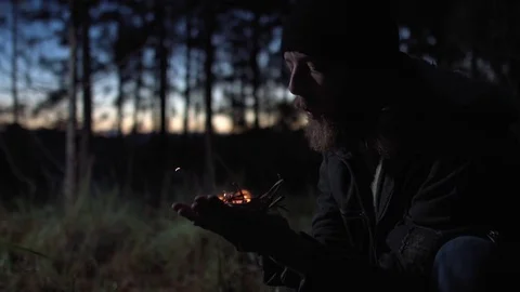 Bearded man lighting a campfire in a forest at dawn. Blowing fire on his hand. Stock Footage 76442472