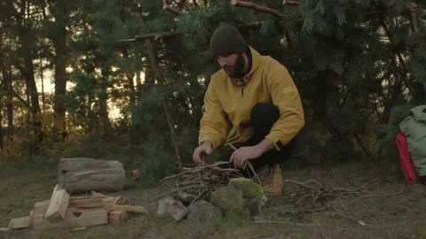 A bearded man lights a fire near a makeshift shelter made of pine branches. Stock Footage 220190406