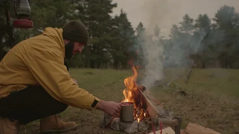 A bearded man lights a fire near a makeshift shelter made of pine branches. Stock Footage 220191574