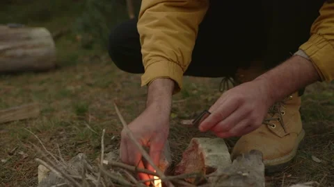 A bearded man lights a fire near a makeshift shelter made of pine branches. Stock Footage 220191613