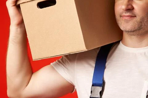 Bearded man in a loader work suit holds a cardboard box on his shoulder Stock Photos