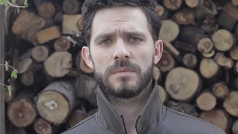 Bearded Man Looking Sad into the Camera with Wooden Logs in Background Stock Footage 239654533