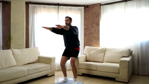 Bearded man in low shape dressed in dark clothes doing squats in living room Stock Footage 151225962