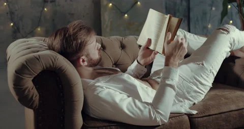 A bearded man is lying on his couch and reading a book Stock Footage 134680580