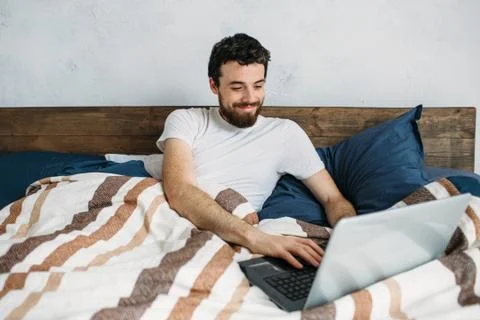 Bearded man lying in morning bed with laptop Stock Photos