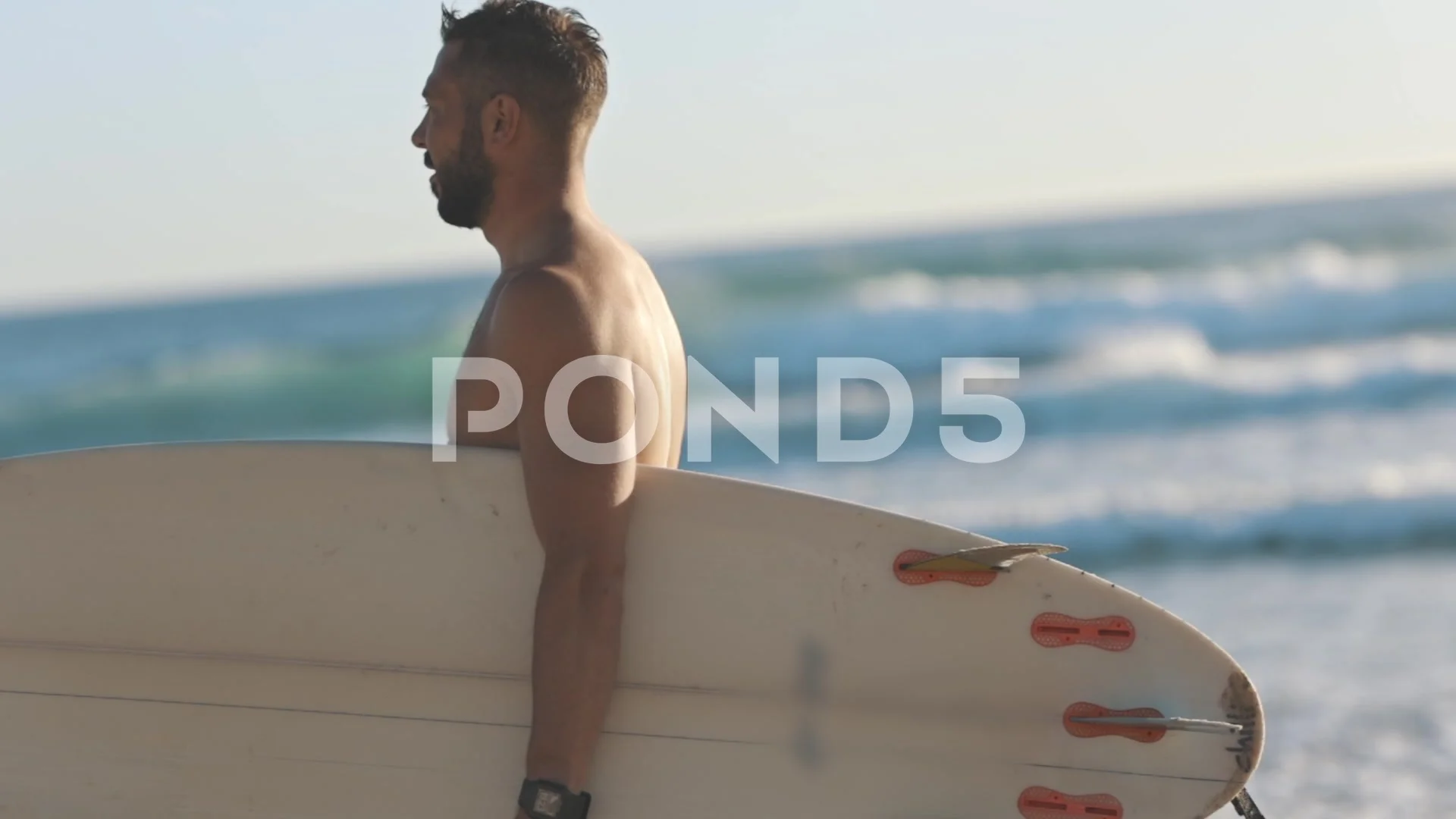 Bearded man with a naked torso walks along the seashore with a surfboard