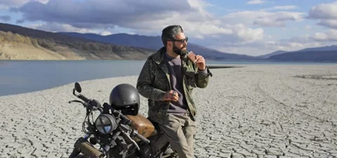 Bearded man on old cafe racer motorcycle near lake, on dried cracked shore Stock Footage 140625323