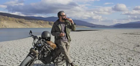 Bearded man on old cafe racer motorcycle near lake, on dried cracked shore Stock Footage 140625327