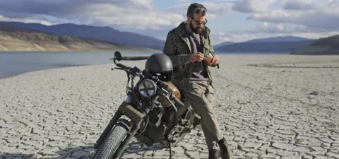 Bearded man on old cafe racer motorcycle near lake, on dried cracked shore Stock Footage 140625360