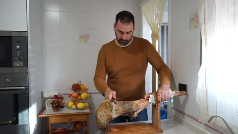 Bearded man in an orange sweater cutting ham in the kitchen at home Stock Footage 149397601