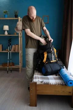 Bearded man packing backpack on bed preparing for hike at home. Travel, hiking Stock Photos