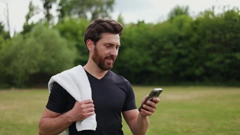 Bearded man passing by grass field while reading news through modern smartphone 스톡 동영상 278022465