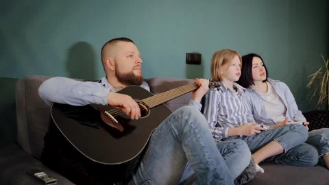 Bearded man playing guitar while his wife and child play video games Stock Footage 265104801