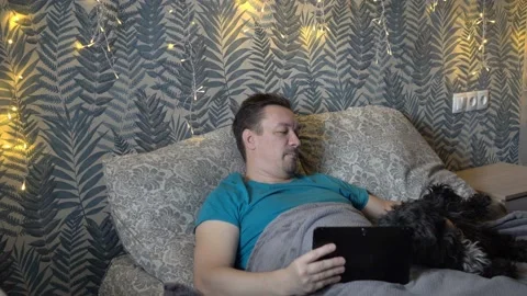 A bearded man plays and strokes a black mongrel dog while lying with a digital Video stock 149441142
