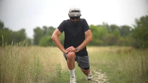 Bearded man pulling knee to chest on grassy trail for outdoor warm-up exercise Video stock 313951512