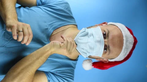 Bearded man in Santa hat puts on a face mask on a blue background, concept of Vidéo 142205055