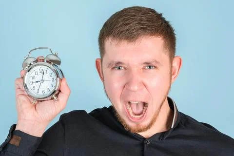 A bearded man screams while holding an alarm clock near his face, close-up Stock Photos
