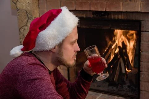 A bearded man sits in front of a lighted fireplace with a glass of red wine. Foto stock