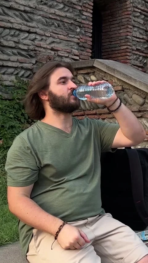 Bearded man sits on wall and drinks water from plastic bottle on old city street Video stock 280854090