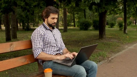Bearded man is sitting at bench in park and working with laprop. He is typing Stock Footage 99739950