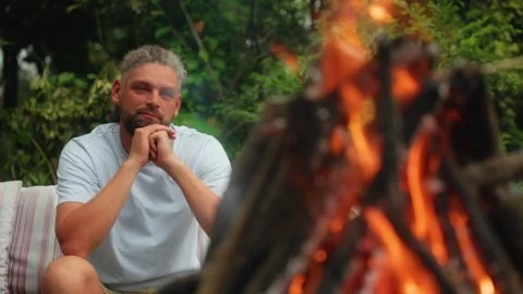 Bearded Man Sitting by Campfire in Garden with Thoughtful Expression. Stock Footage 338367965