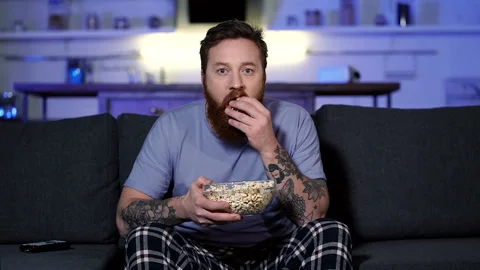 Bearded man sitting on the sofa, eating popcorn and watching tv. Stock Footage 122201137