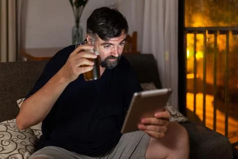 Bearded man on sofa uses digital tablet and drink a beer in dining room, nigh Stock Photos