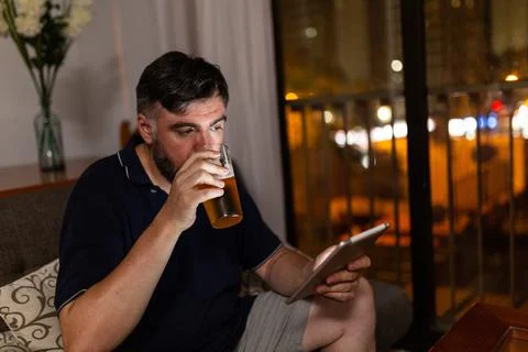 Bearded man on sofa uses digital tablet and drink a beer in dining room, night Stock Photos