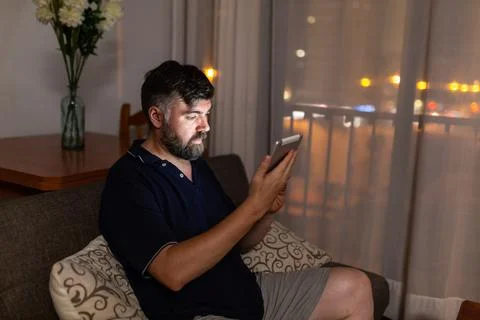 Bearded man on sofa uses digital tablet in dining room overnight Stock Photos