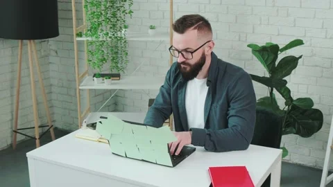 Bearded man types at the laptop and write notice on the paper notebook. Stock Footage 155403973