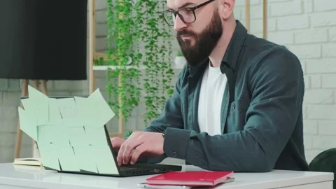Bearded man types at the laptop and write notice on the paper notebook. Stock Footage 155641006