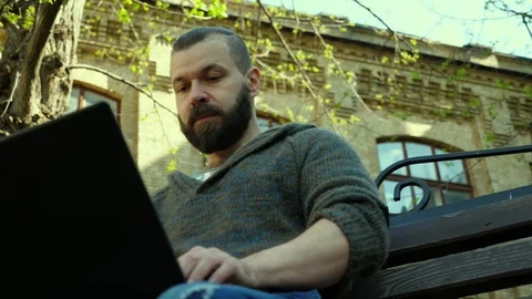 The bearded man typing on a laptop outside on a bench and seeks inspiration Stock Footage 88782795