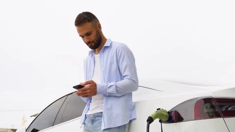 Bearded man uses a mobile application to control the battery of an electric car 스톡 동영상 251764124