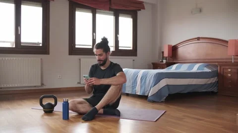 Bearded man using his mobile phone while resting on the floor. Home exercise. Stock Footage 158488001