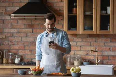 Bearded man using smartphone, distracted from cooking in kitchen Foto stock