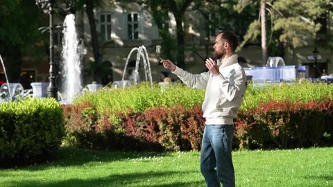 Bearded Man Using Smartphone on Selfie Stick Near Subotica Park Fountain Видео 334615804