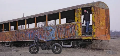 Bearded man walks through old graffiti train, near old cafe racer motorcycle Stock Footage 140626585