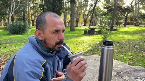 A bearded man wearing a blue jacket drinks mate at a wooden table surrounded by Stock Footage 314876767