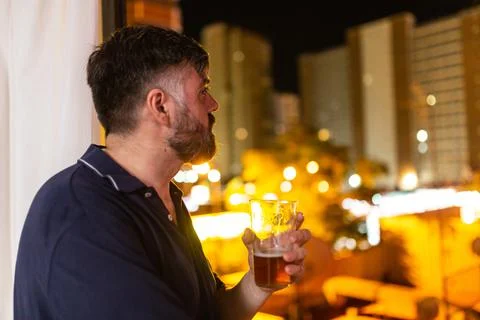 Bearded man by the window drinking beer at night, dressed in polo shirt Stock Photos
