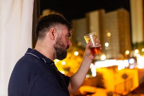 Bearded man by the window drinking beer at night, dressed in polo shirt Stock Photos
