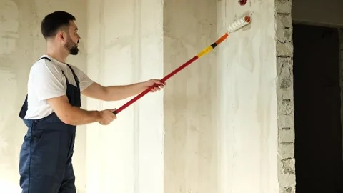 A bearded man in a work uniform is priming the walls after applying gypsum Stock Footage 318896961