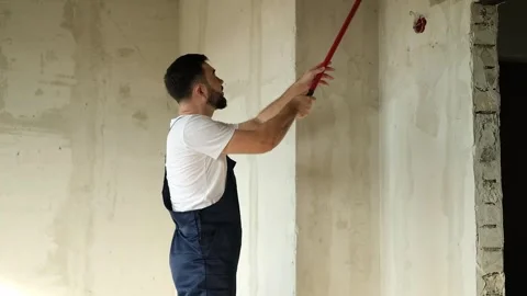 A bearded man in a work uniform is priming the walls after applying gypsum Stockbeeldmateriaal 346689560