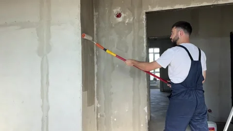 A bearded man in a work uniform is priming the walls after applying gypsum Stock Footage 346689692