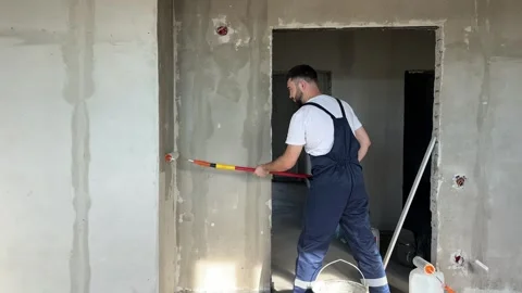 A bearded man in a work uniform is priming the walls after applying gypsum Stock Footage 346691586
