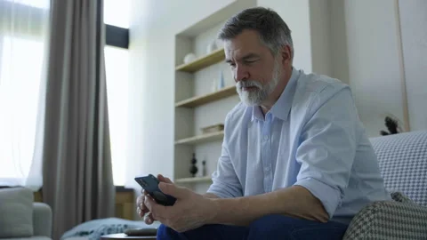 Bearded mature man sitting on sofa use mobile phone smiles at home Video stock 244073442